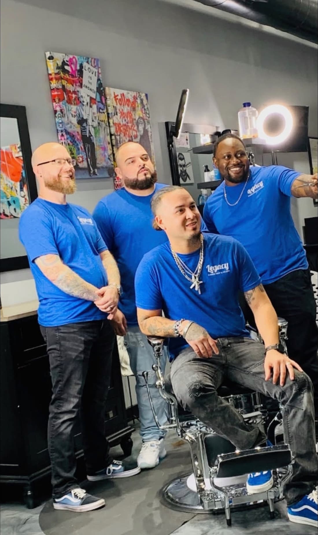 Four Legacy barbers in matching blue Legacy shirts posing around a barber chair in the shop